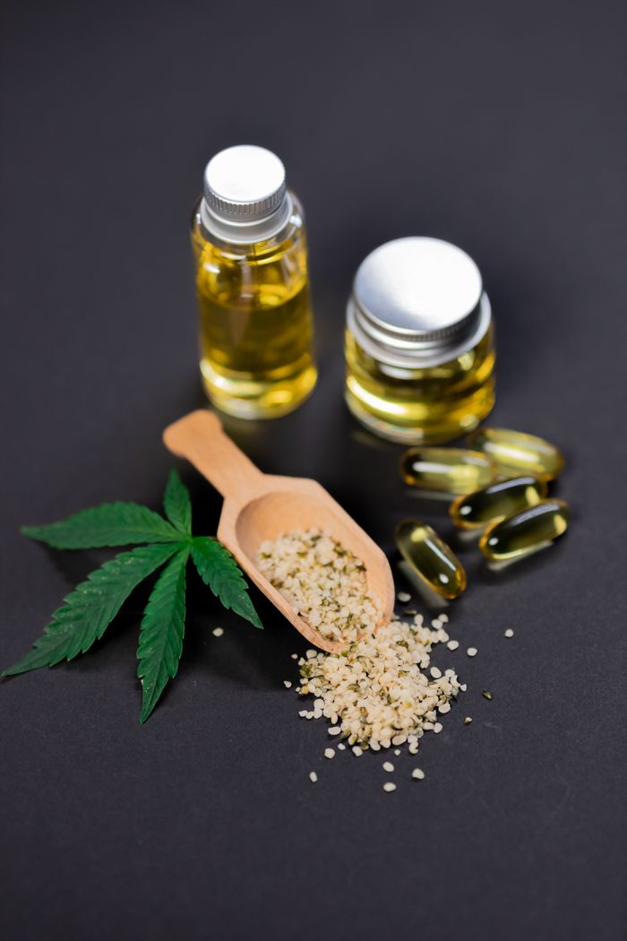 Flat lay of cannabis oil, hemp seeds, capsules, and leaf on dark background.